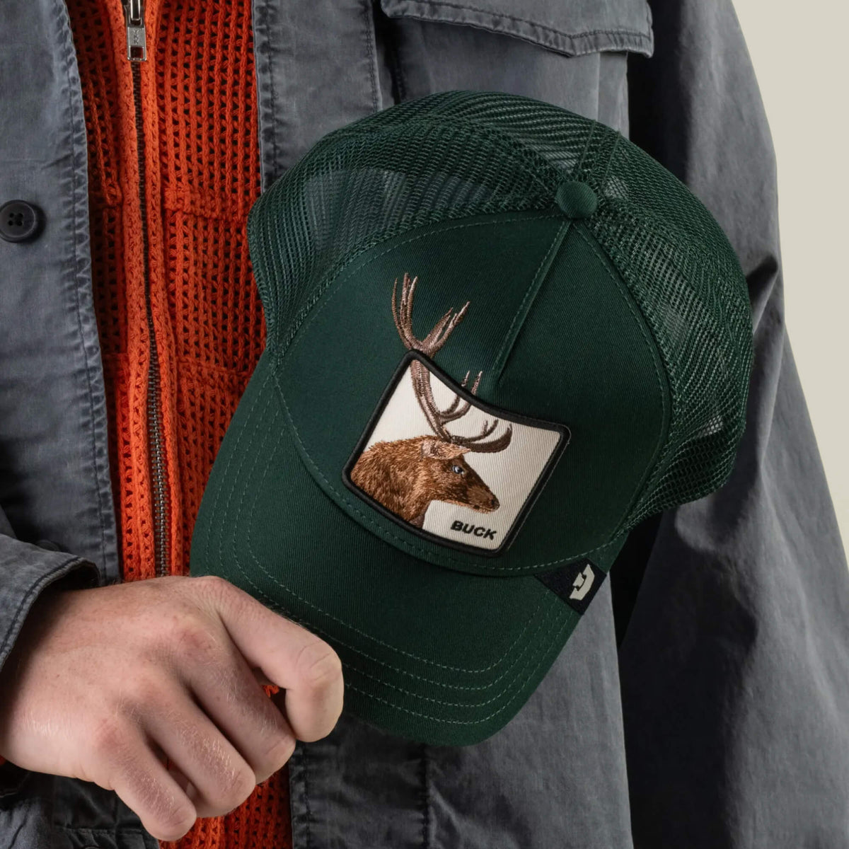 A person holds the Goorin Bros. Over the Line Buck dark green trucker hat, featuring an embroidered patch with a buck and antlers and breathable mesh panels, while wearing an orange sweater and a gray jacket.