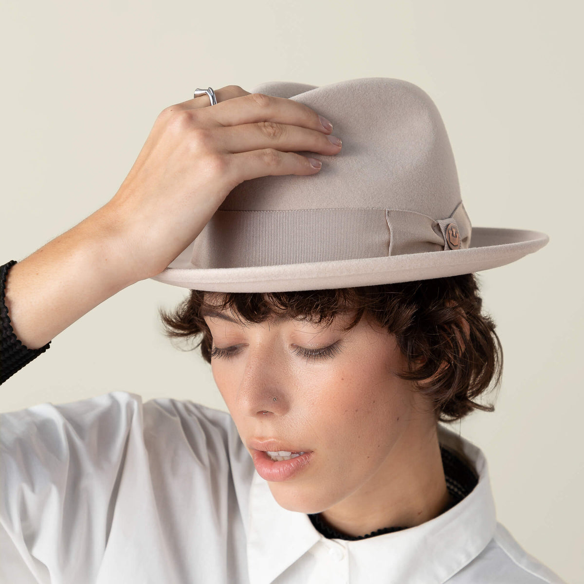 A person with short brown hair wears the Dean the Butcher fedora by Goorin Bros., featuring a grosgrain hatband, and a white collared shirt. They hold the brim and look downward with closed eyes.