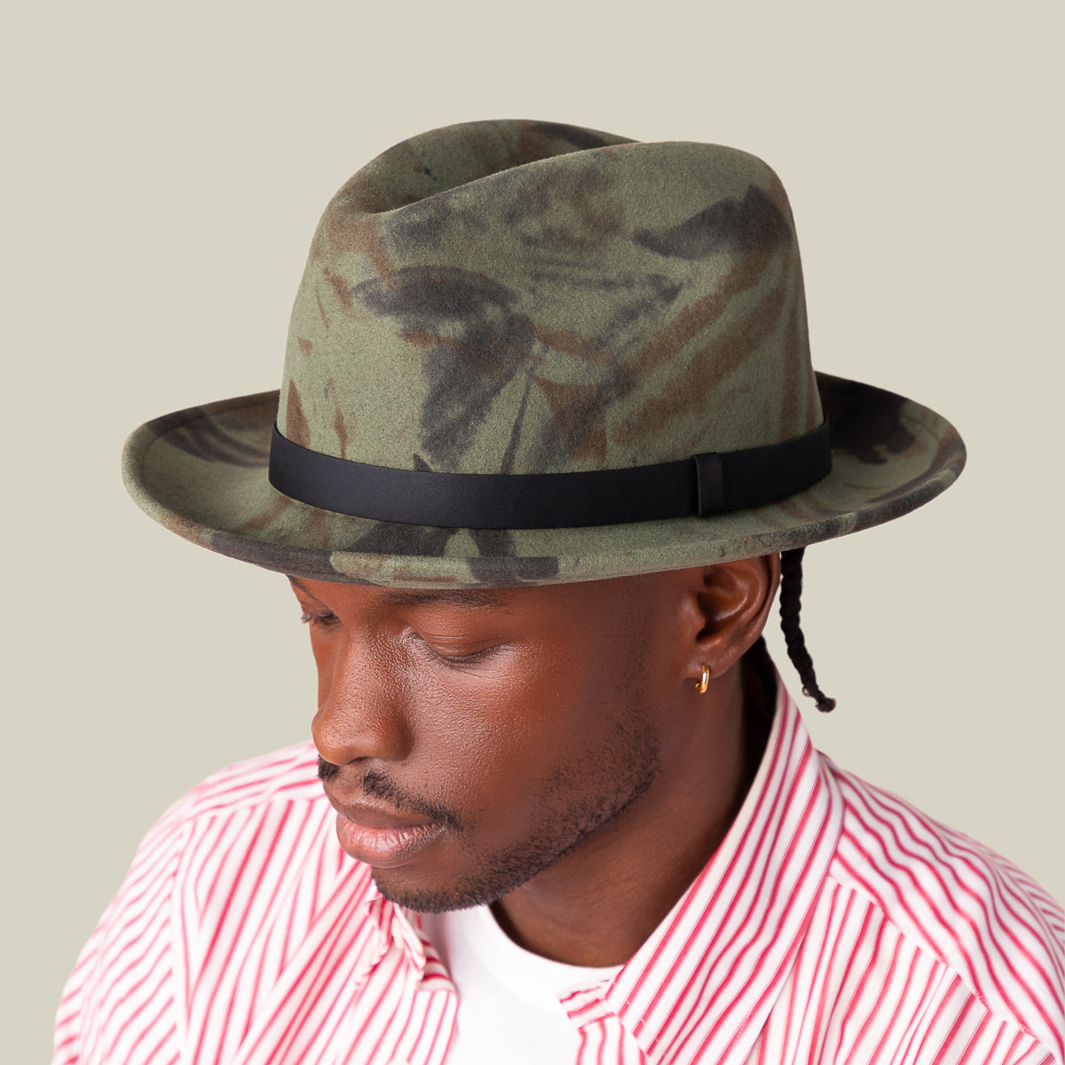 A man wearing Goorin Bros.' Dean the Butcher fedora with a green camo pattern and grosgrain hatband, a gold hoop earring, and a red and white striped shirt looks downward against a plain beige background.