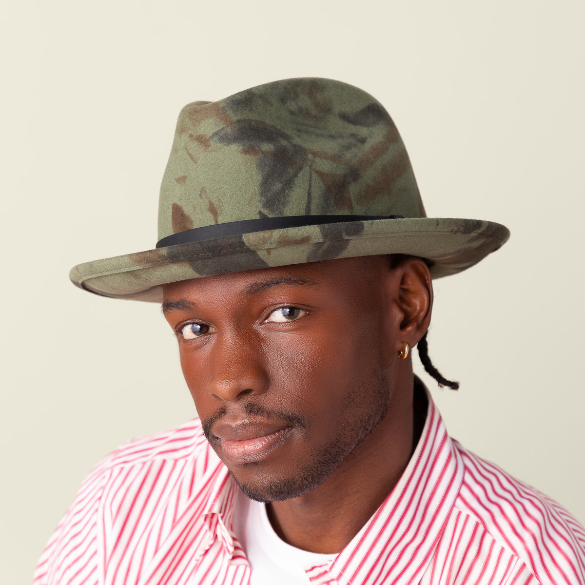 A man wearing the Dean the Butcher fedora by Goorin Bros., featuring a green camouflage pattern, pairs it with a red and white striped shirt and a small gold hoop earring, gazing at the camera against a plain background.