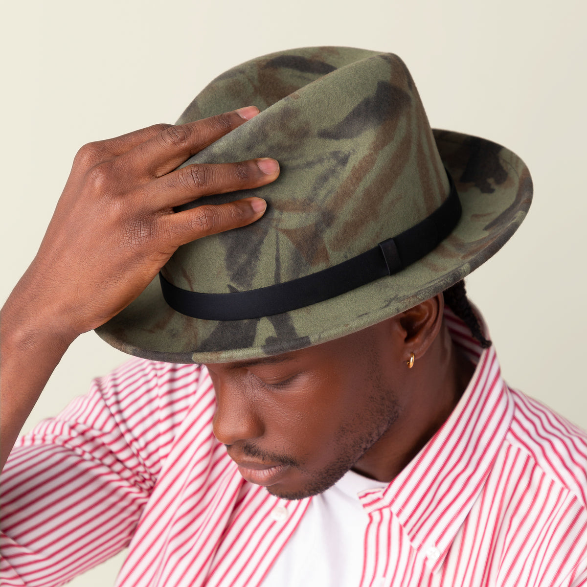 A man wearing the Goorin Bros. "Dean the Butcher" fedora with a grosgrain hatband and a red-and-white striped shirt looks down while holding the brim of his hat.
