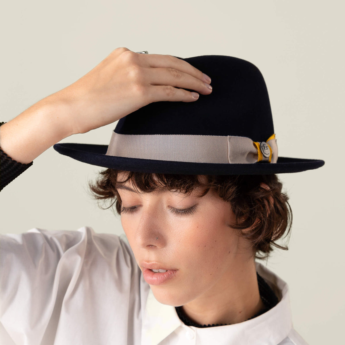 A person wearing the Goorin Bros. "Dean the Butcher" fedora with a black grosgrain hatband featuring a gray ribbon and yellow detail, tilting the brim with one hand and dressed in a white shirt.