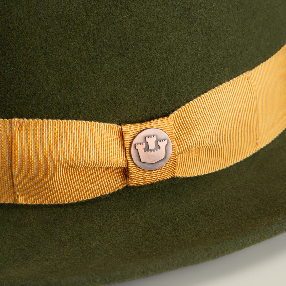 A close-up of Dean the Butcher by Goorin Bros., a green American-made fedora with a yellow grosgrain hatband and a round metal pin featuring a castle emblem at its center.