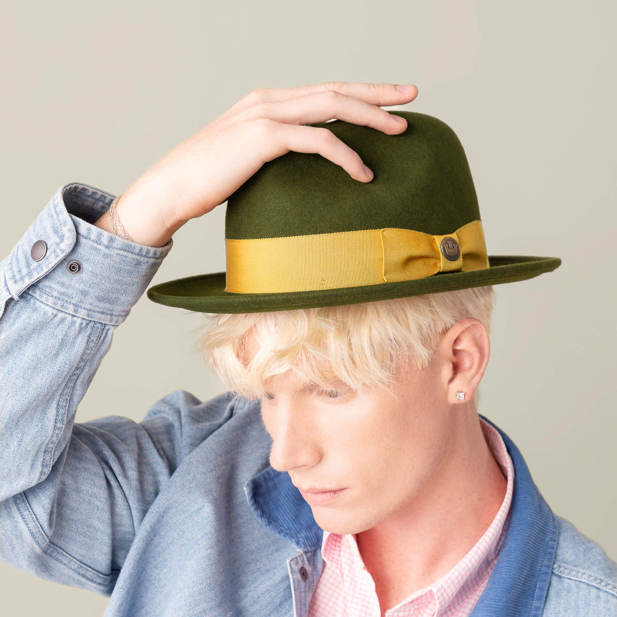 A person with light hair wears the Dean the Butcher hat by Goorin Bros.—a green all-season hat with a yellow grosgrain band—paired with a light denim jacket and pink shirt, holding the brim and looking down.