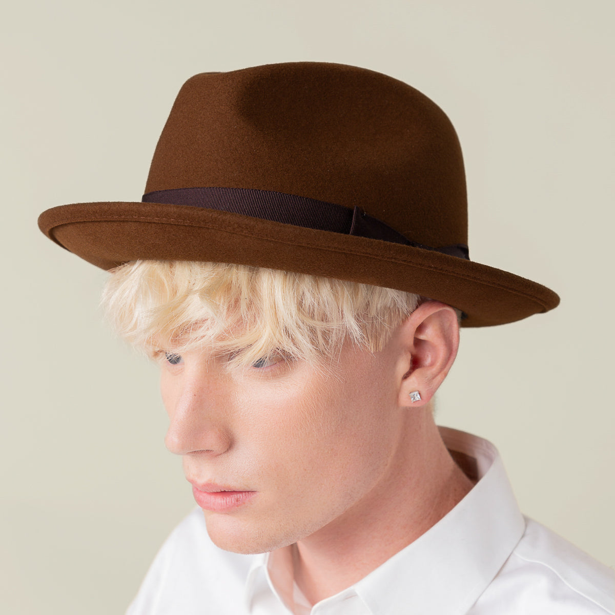 A person with light blonde hair faces left, wearing the Goorin Bros. Dean the Butcher fedora with a grosgrain hatband and a white collared shirt. They have a stud earring and a neutral expression.
