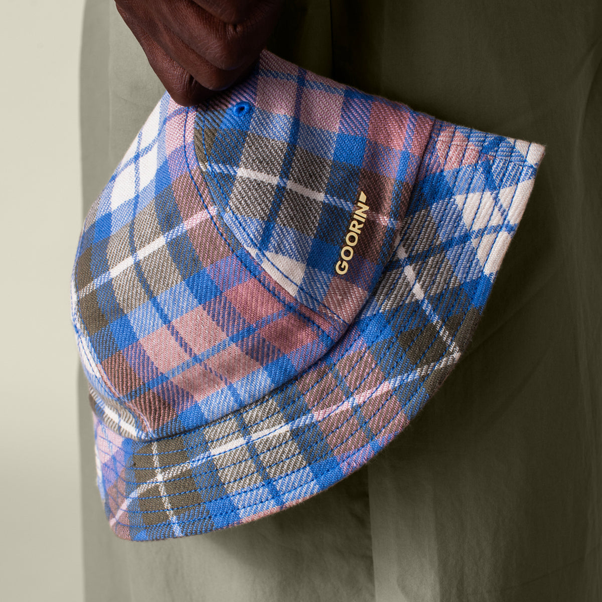 A hand holds the Bespoke Wordmark Bucket by Goorin Bros., a cotton linen bucket hat featuring blue, pink, white, and brown plaid patterns with the GOORIN wordmark embroidered in gold on the side.