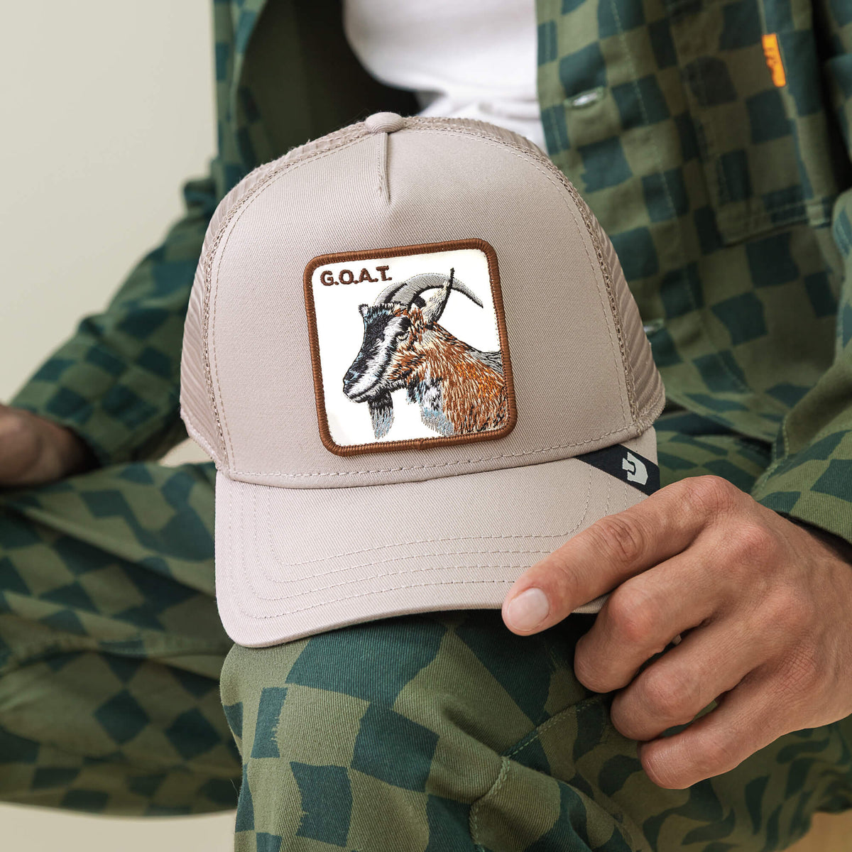 A person holds the Goat Trucker hat by Goorin Bros., a beige trucker featuring a bold illustrated goat patch with "G.O.A.T" lettering, paired with green patterned neutral-toned clothing.