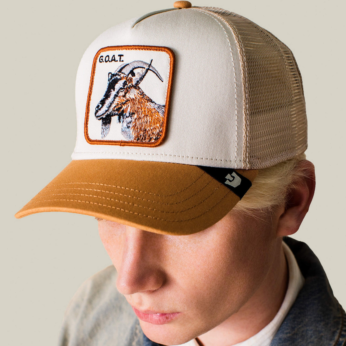 A person wears the Goorin Bros. Goat Trucker hat in tan and white neutrals, featuring a bold front patch with an embroidered goat and "G.O.A.T." letters.