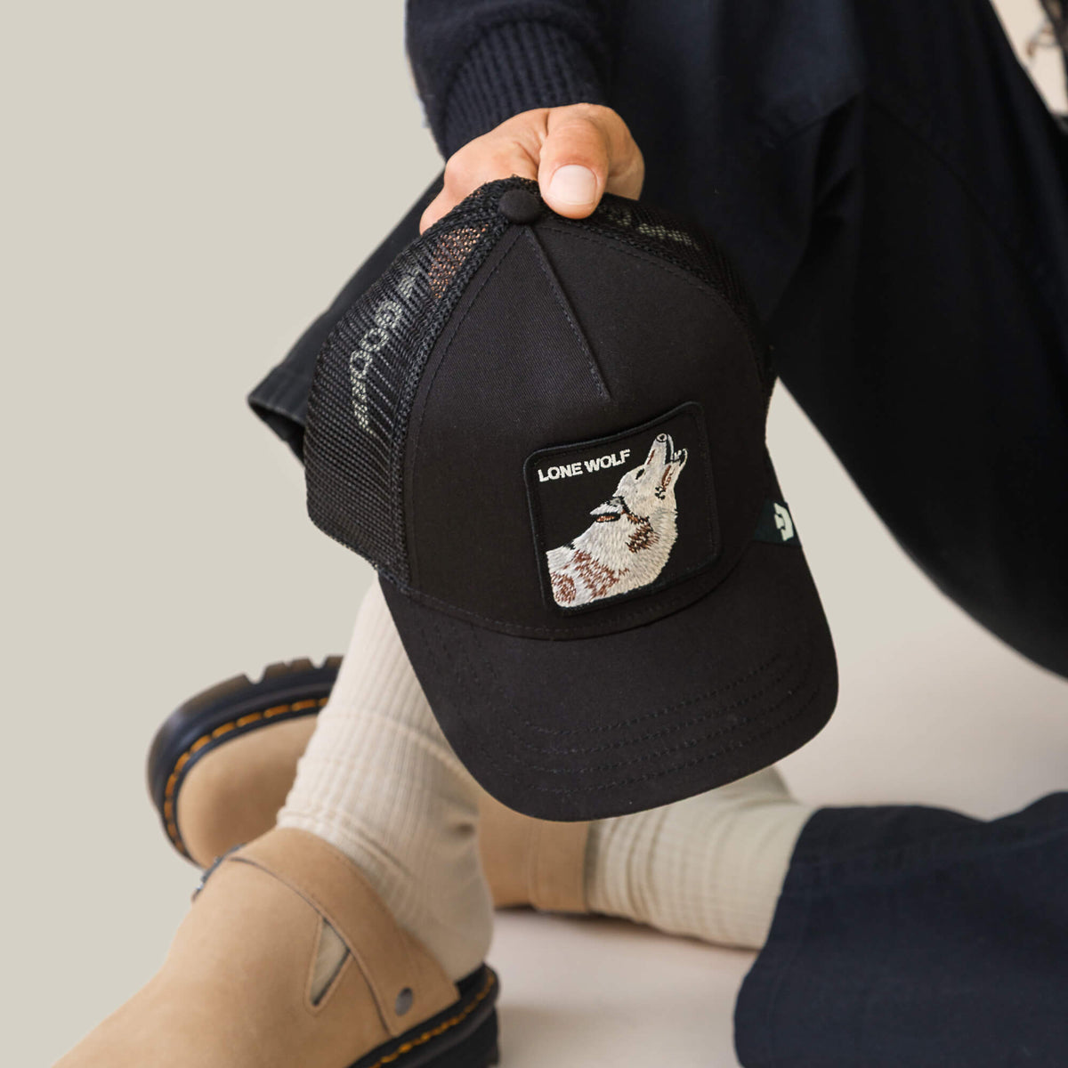 A person wears the Goorin Bros. Lone Wolf Trucker, a black mesh cap with a patch showing a wolf and "Lone Wolf" text, paired with beige socks, sandals, and dark pants for an effortlessly cool, rebellious style.