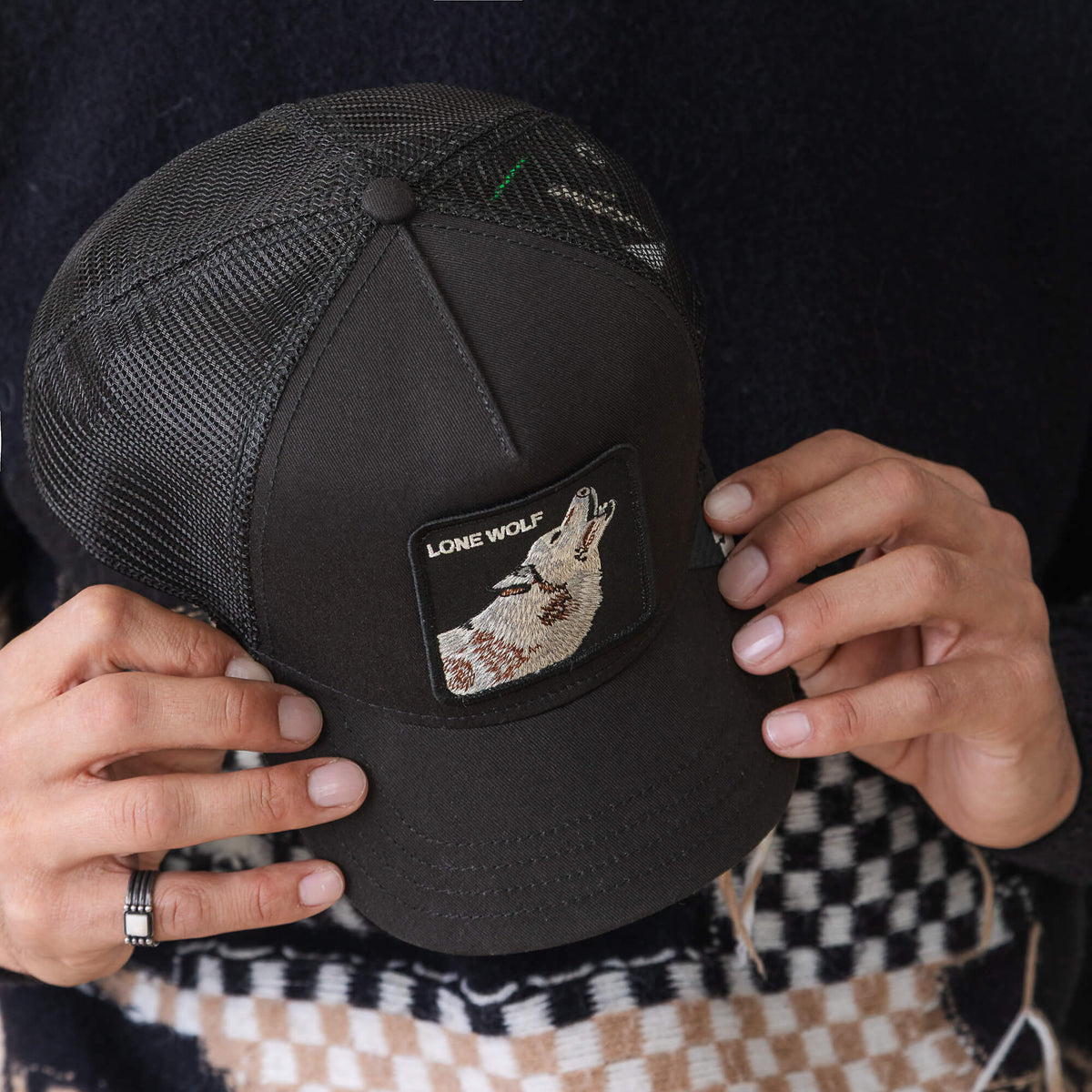 Person holding the Goorin Bros. Lone Wolf Trucker, a black mesh hat with an embroidered patch of a howling wolf and "LONE WOLF" on the front.