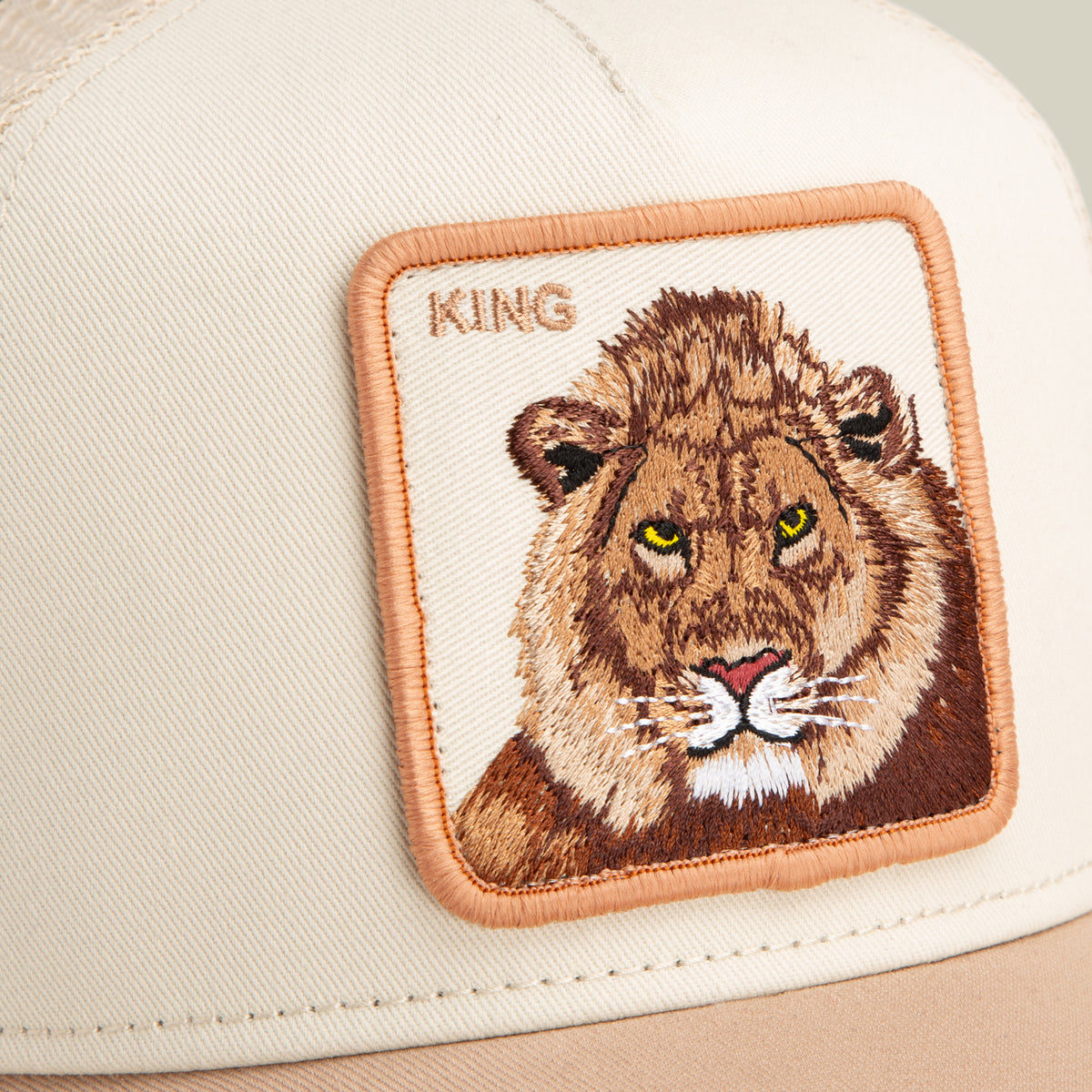 Close-up of the Goorin Bros. King Trucker cap in beige, featuring an embroidered lion patch with "KING" stitched above it.