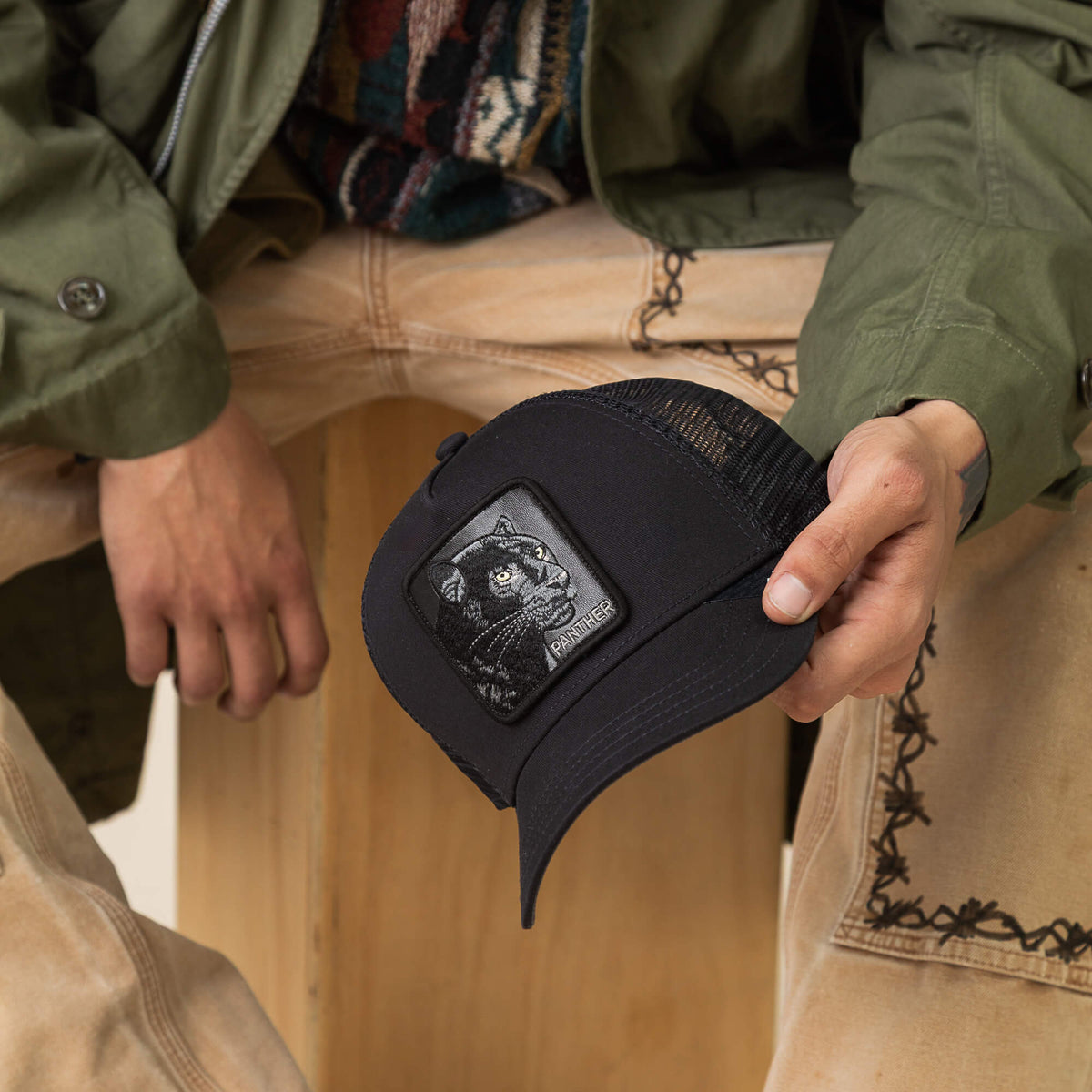 A person holds the Goorin Bros. Panther Trucker hat, featuring a black cat patch, while sporting an olive jacket and light-colored pants with decorative stitching.