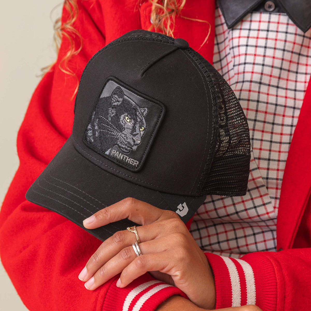 A person in a red jacket holds the Goorin Bros. Panther Trucker—a black mesh cap with a patch featuring a black panther illustration and "PANTHER" on the front.