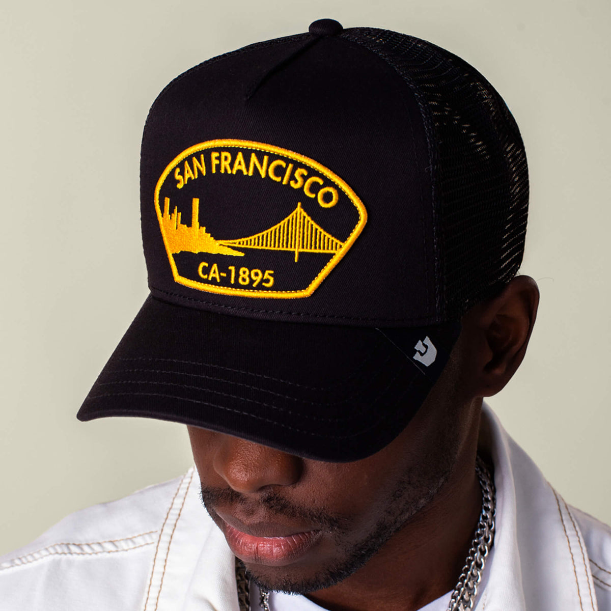 A person wearing a black mesh Goorin Bros. San Francisco hat with a Golden Gate patch and a white jacket, looking down.