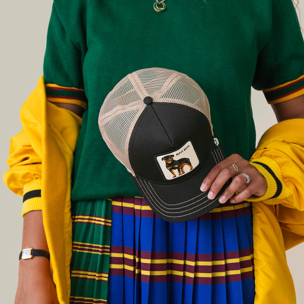 Wearing a green top, blue striped skirt, and yellow jacket, they show off rings and the Goorin Bros. Bad Boy Trucker cap featuring a dog patch for a bold, edgy style.