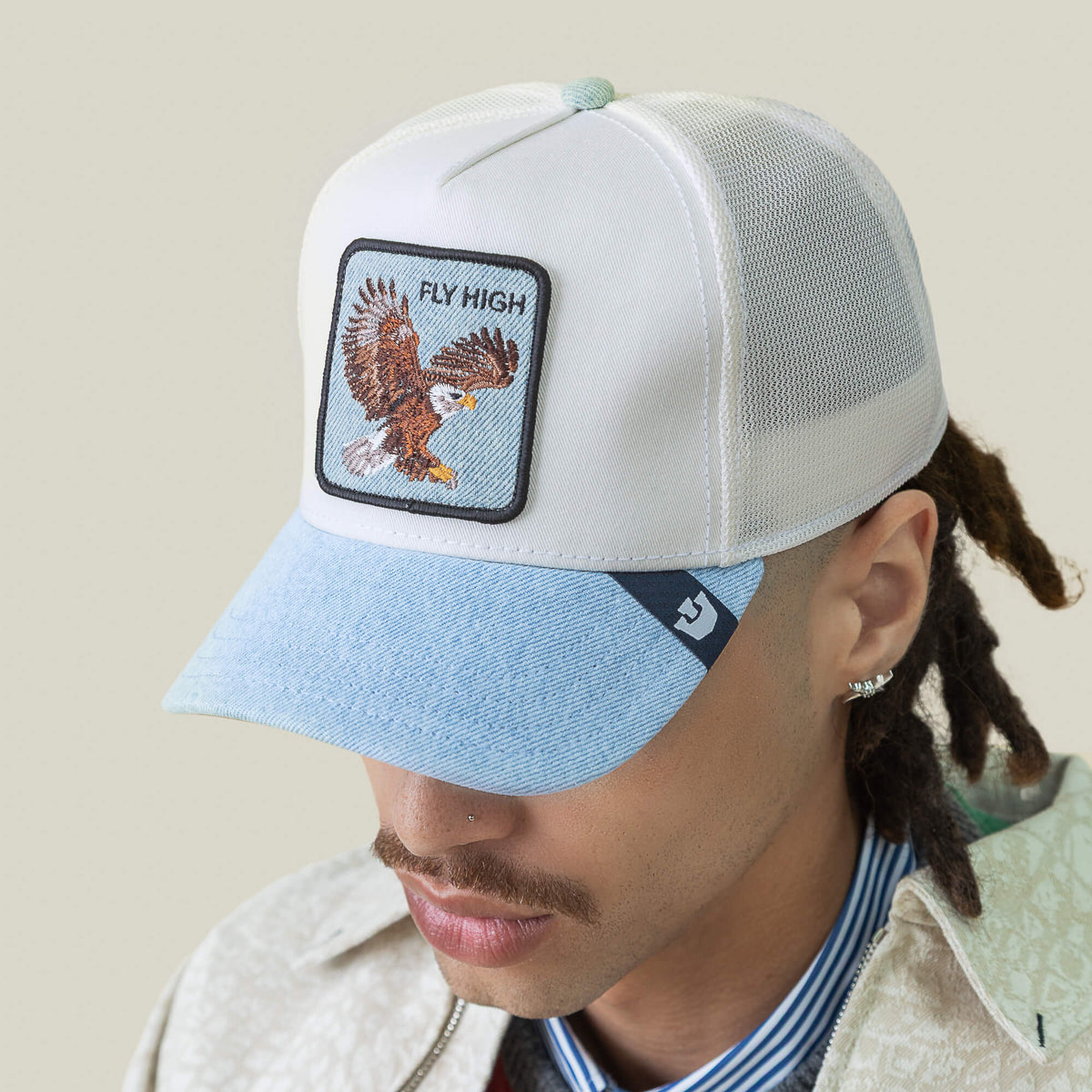 A person sporting the Goorin Bros. Denim Fly High cap—light mesh, blue brim, eagle patch with "FLY HIGH"—plus a mustache, earrings, and dreadlocks: true style for altitude junkies chasing pure elevation.