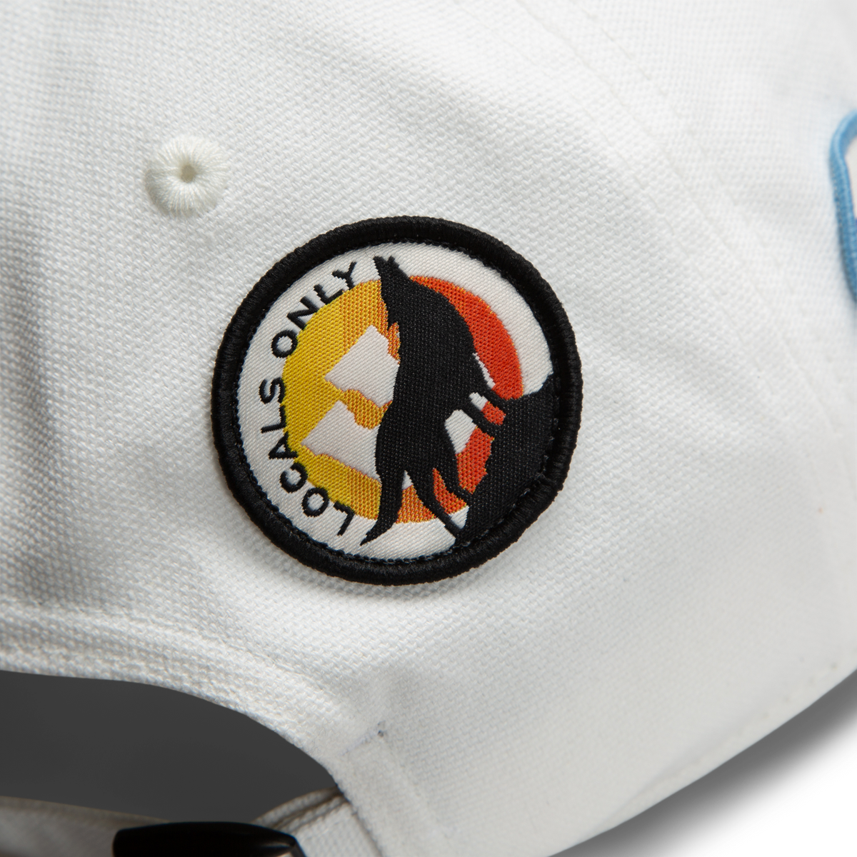 A close-up of the Goorin Bros. Pop Appeal white cap, featuring a LOCALS ONLY patch with a black wolf on an orange-yellow background, stylishly paired with white canvas sneakers and collectible badges.