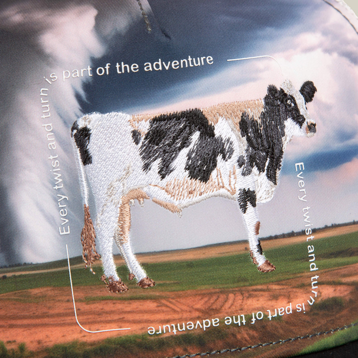 The "Through the Storm - Cow" trucker cap by Goorin Bros. features an embroidered cow patch with the phrase “Every twist and turn is part of the adventure” set over a rural landscape print on a black and white design.