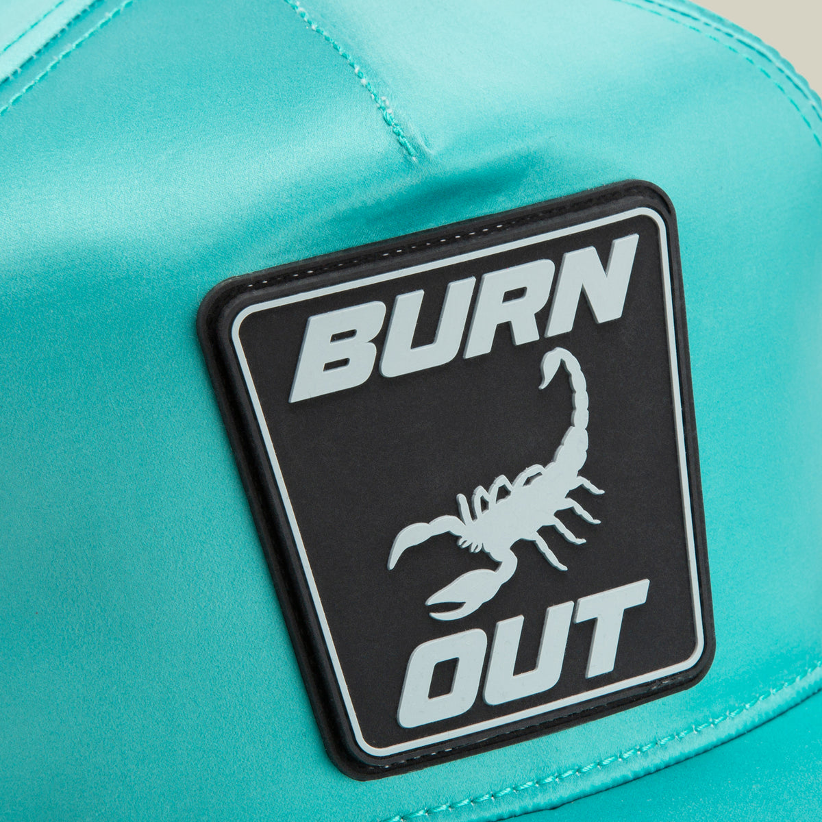 Close-up of the Goorin Bros. Pole Position - Road Kill racing cap in teal, featuring sleek chrome satin stripes and a black and white patch displaying the words BURN OUT with a scorpion image.