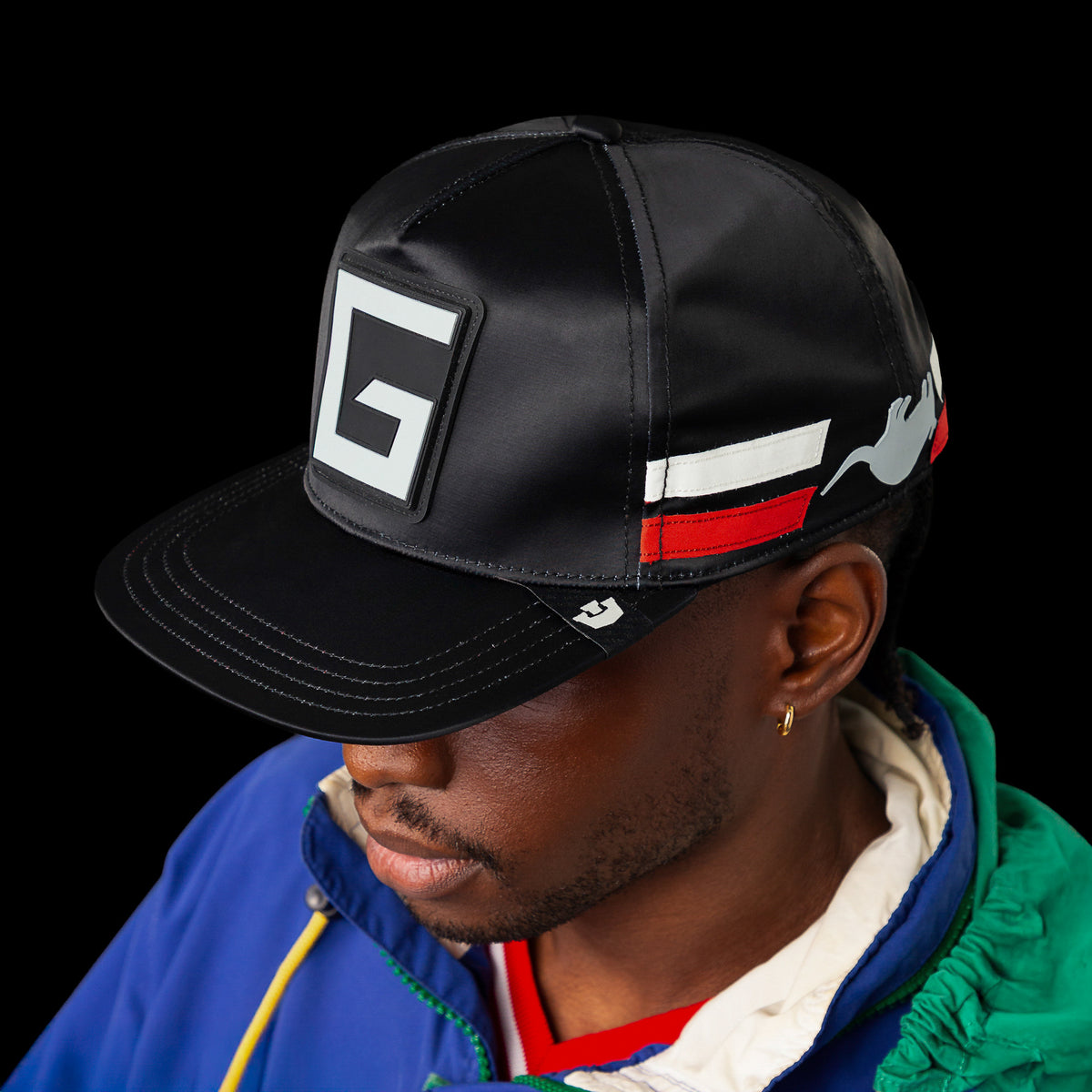 A person wearing the Goorin Bros. “Pole Position - Road Kill” hat, featuring a large G logo and bold white and red chrome satin stripes, paired with a blue jacket with a white hood, posed against a black background.