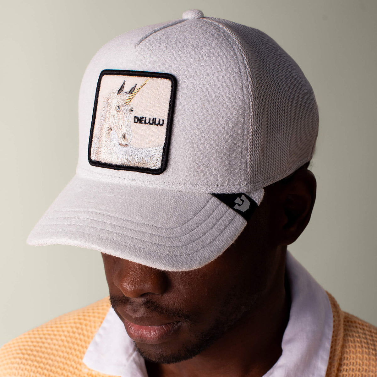 A man wears the Goorin Bros. Terry Cloth Unicorn cap, light gray with a unicorn patch reading DELUU, paired with a beige terry cloth jacket over a white collared shirt.