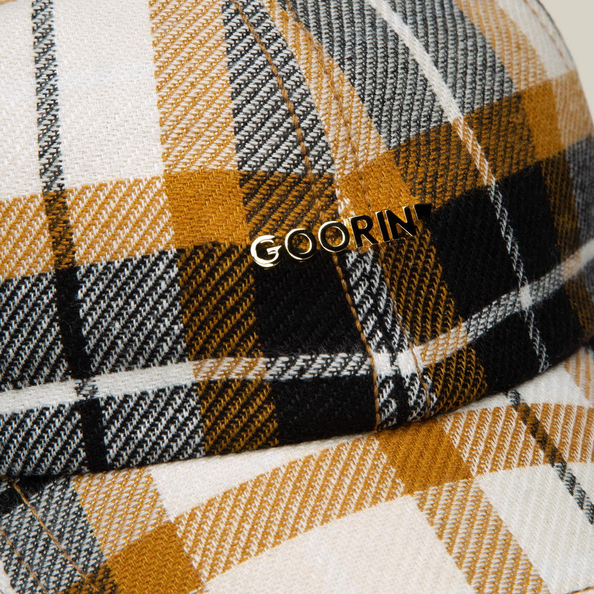 Close-up of the Bespoke Wordmark V2 plaid cap in yellow, black, and white by Goorin Bros., featuring a metallic brand logo—a stylish cap for any wardrobe.