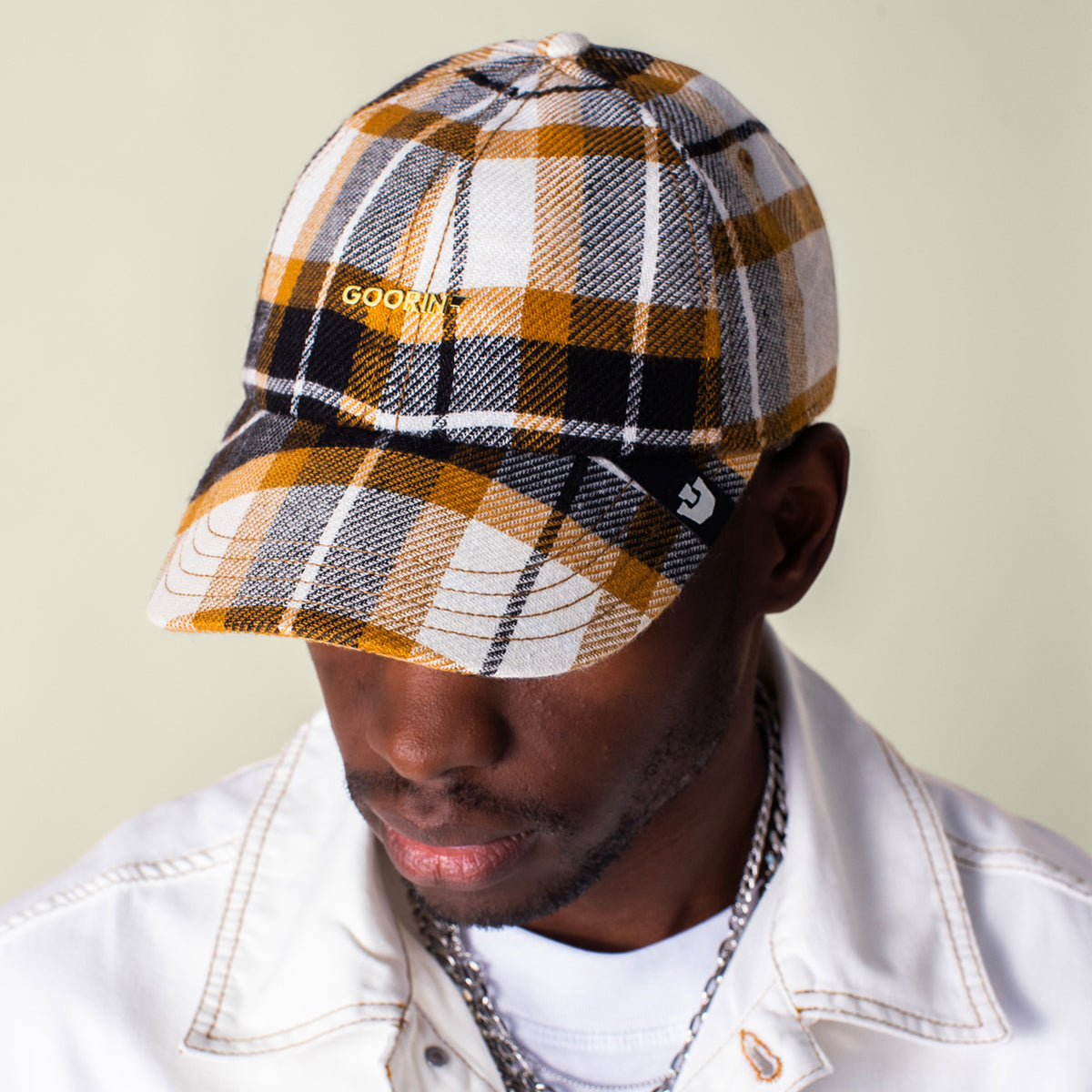 A man wearing the Bespoke Wordmark V2 plaid cap by Goorin Bros. and a white jacket looks down against a plain background.