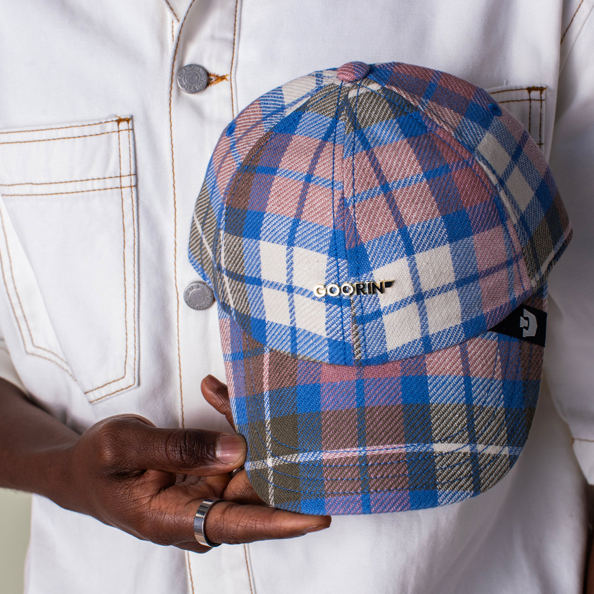 A person in a white jacket holds a Bespoke Wordmark V2 plaid cap by Goorin Bros., showcasing the stylish cotton linen papa cap design with the distinctive GOORIN branding on the front.