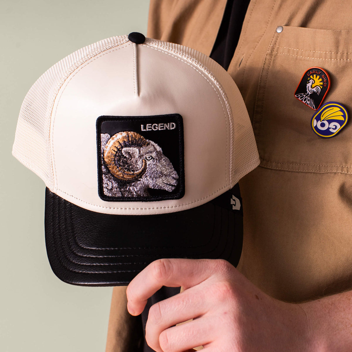A person holds the beige and black Goorin Bros. Faux Leather Ram cap labeled LEGEND. They wear a tan shirt with two colorful pins on the pocket.