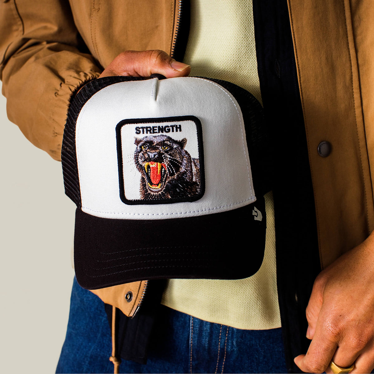 A person holds the Goorin Bros. Strength Panther Trucker, a black and white cap featuring a bold panther patch and "STRENGTH," symbolizing power and resilience.
