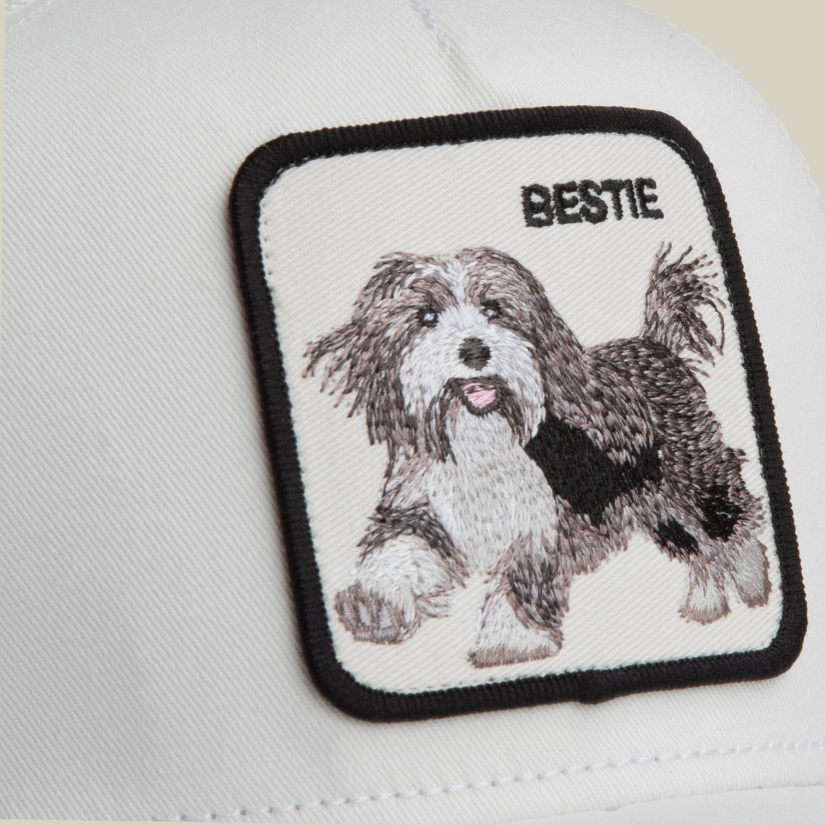 Close-up of the Goorin Bros. Bestie Trucker hat, white fabric with a square embroidered black and white dog patch labeled "BESTIE" in the top right corner.