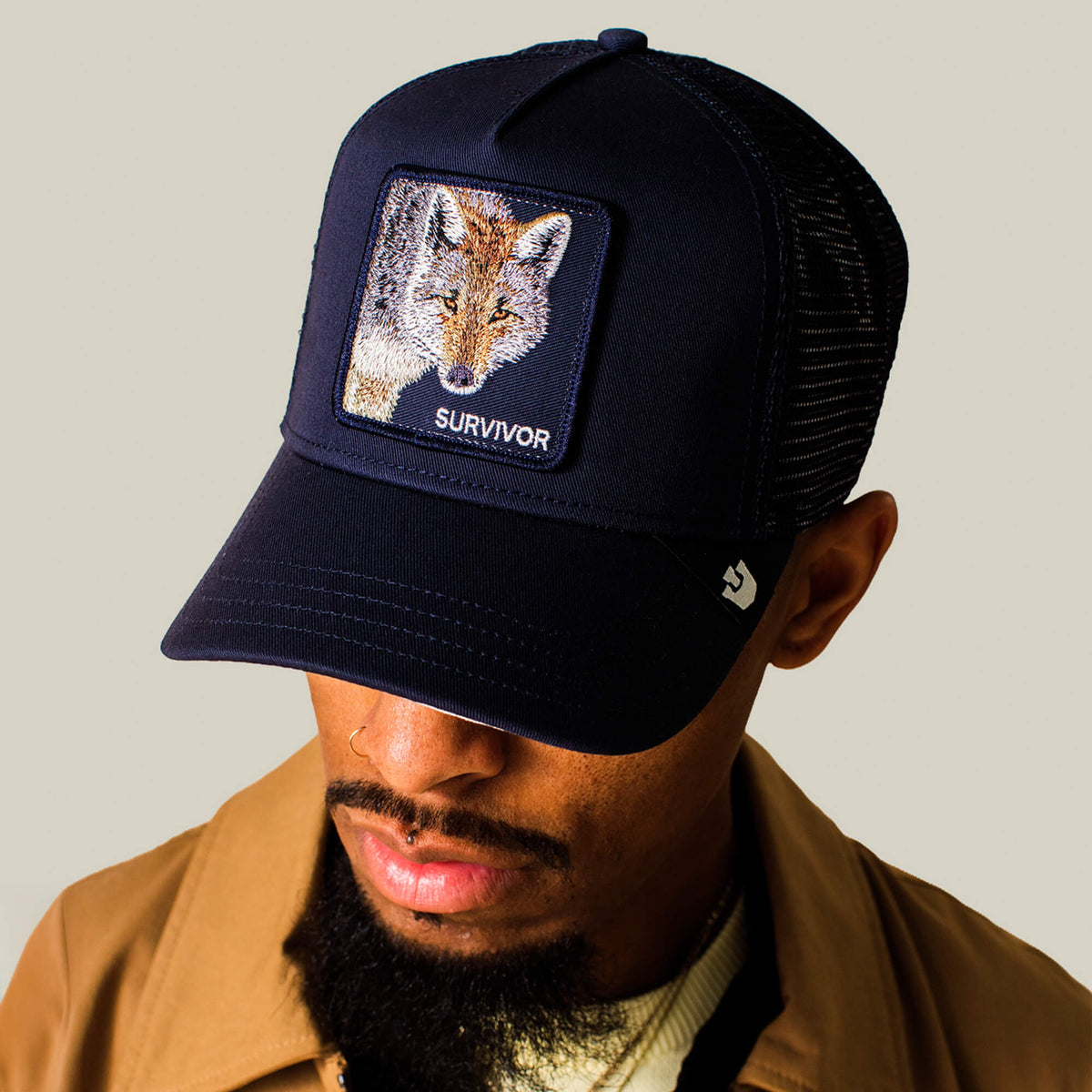 A man wears the Goorin Bros. Mirage Survivor black mesh baseball cap featuring a fox patch labeled “SURVIVOR,” paired with a brown jacket and looking down against a plain beige background, capturing the rugged appeal of this signature hat.