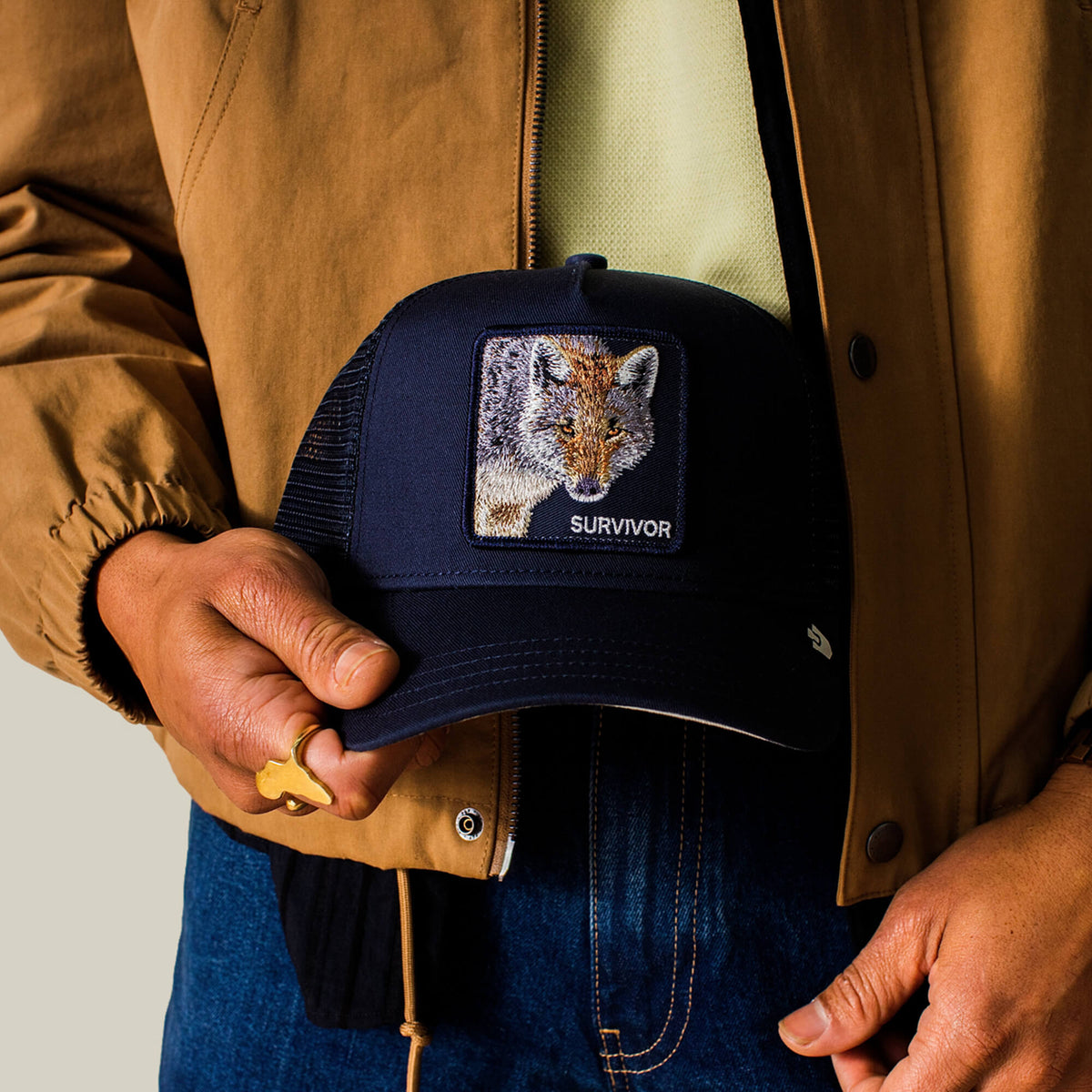 A person in a brown jacket holds the Mirage Survivor navy trucker hat by Goorin Bros., featuring a patch with a fox and the word SURVIVOR.