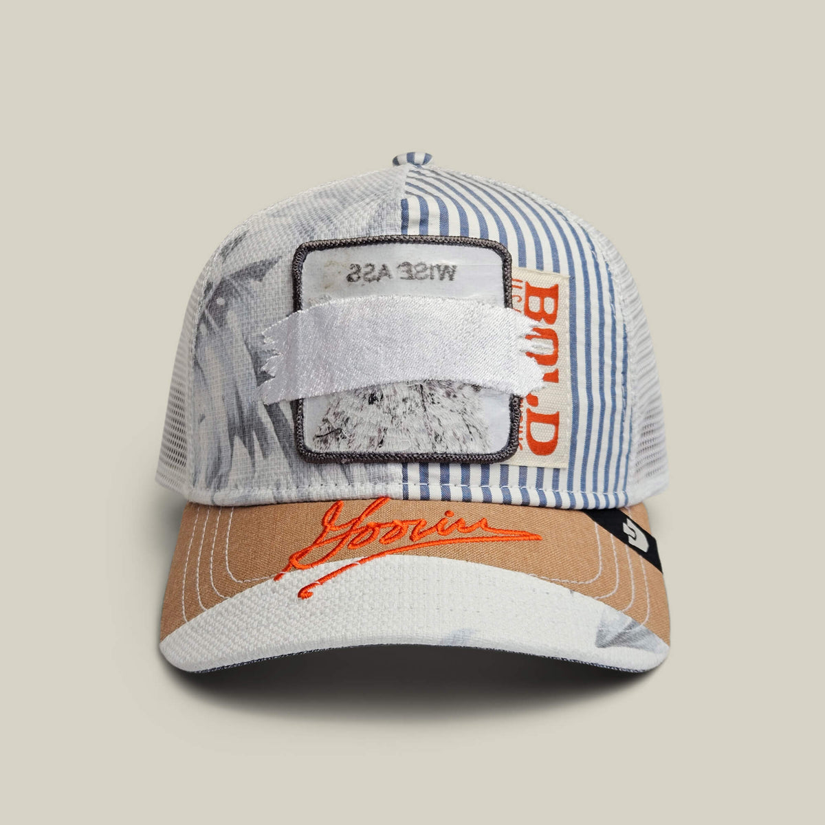 The Goorin Bros. Goorin Rework Trucker in AIR is a limited edition multicolored cap featuring blue stripes, orange text, a brown brim, and a white patch with WISE ASS partially covered plus red embroidered branding on the front.