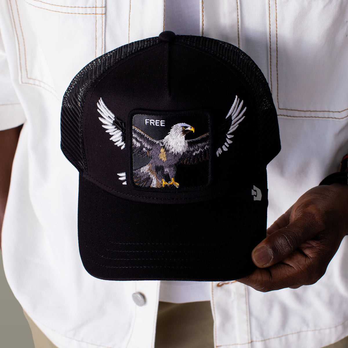 A person holds the Goorin Bros. Wings Free hat, a classic black trucker cap with breathable mesh back, featuring an embroidered eagle with outstretched wings and "FREE" above it.