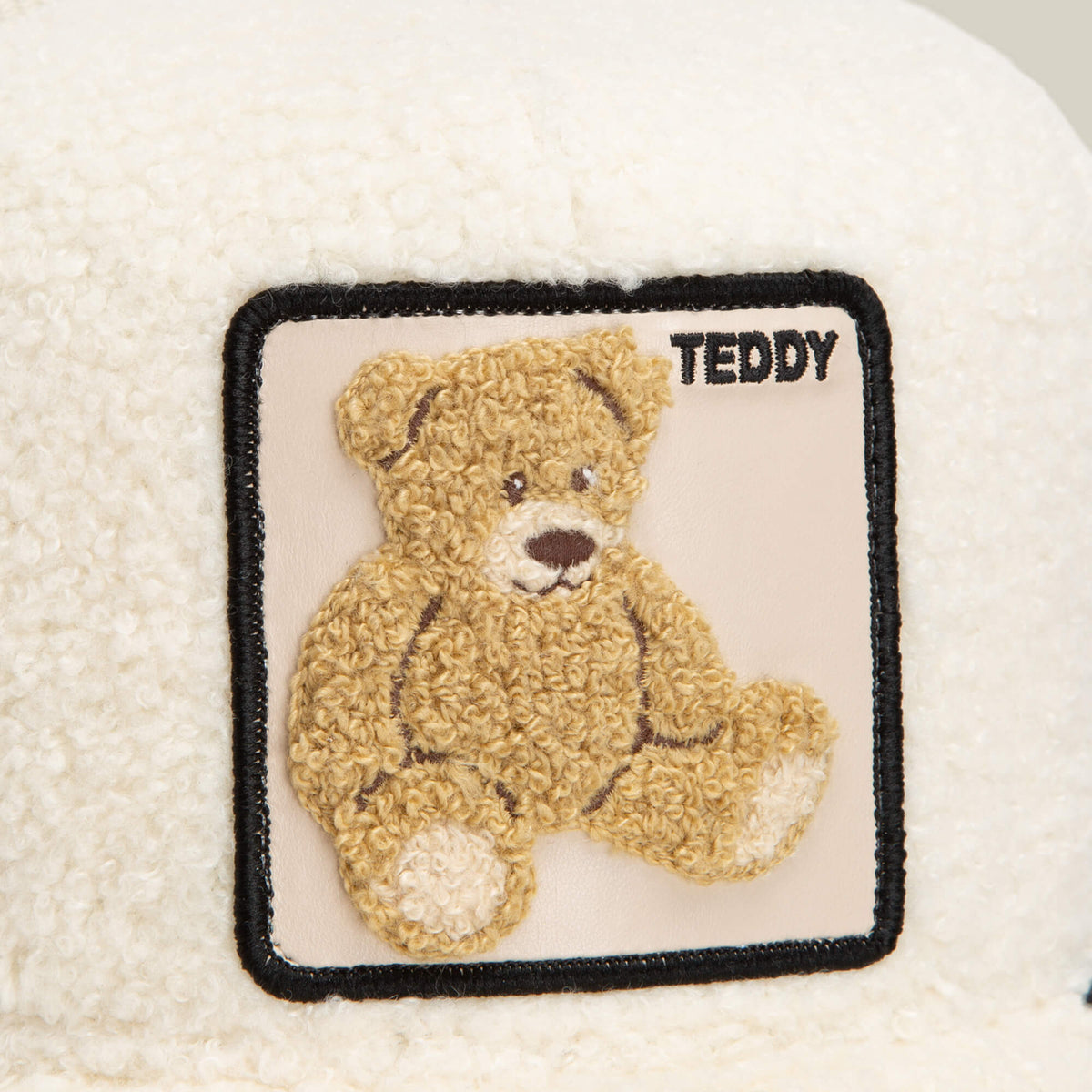 The Double Stuffed - Teddy trucker hat by Goorin Bros. features a cream-colored sherpa fabric adorned with a square patch on the top right, showcasing an embroidered brown teddy bear and the word "TEDDY" in black text, making this cozy accessory both charming and stylish.