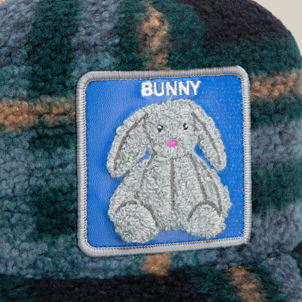 A close-up of the Goorin Bros. Double Stuffed - Bunny trucker hat shows off its blue and green textured design, highlighted by a plush chenille patch featuring a gray bunny and the word "BUNNY" above it on a blue background, and finished with an adjustable snapback for the perfect fit.