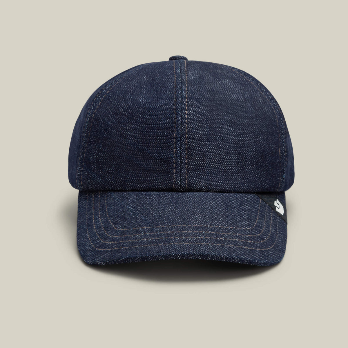 The Noisy Tailored Baseball cap by Goorin Bros. is featured in indigo denim with visible stitching, set against a plain beige background to emphasize its color-driven style and understated confidence.