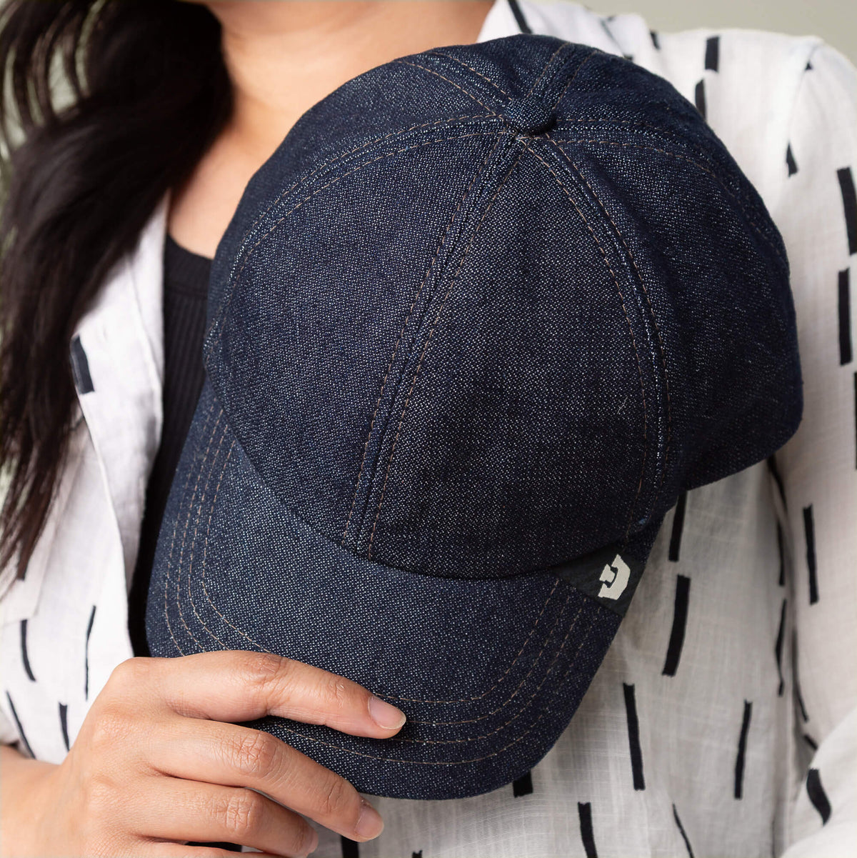 A person holds the Noisy Tailored Baseball cap by Goorin Bros. in dark blue denim, paired with a white shirt featuring black stripes, embodying subtle confidence and color-driven style.