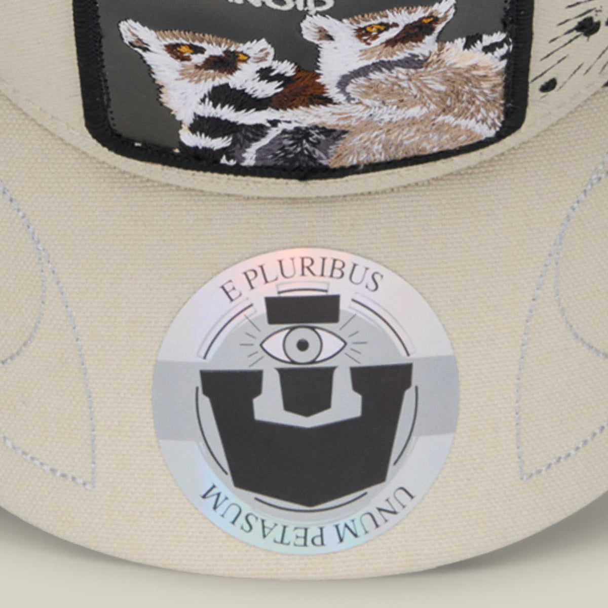 The Goorin Bros. G-Anon is a cream and black trucker hat featuring embroidered raccoon design on the crown and a circular badge with "E Pluribus" text and an all-seeing eye symbol on the front.