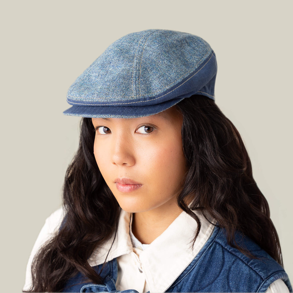 A woman with long dark hair faces forward against a beige background, wearing the Goorin Bros. Full Wattage blue tweed flatcap with a white collared shirt and blue vest.