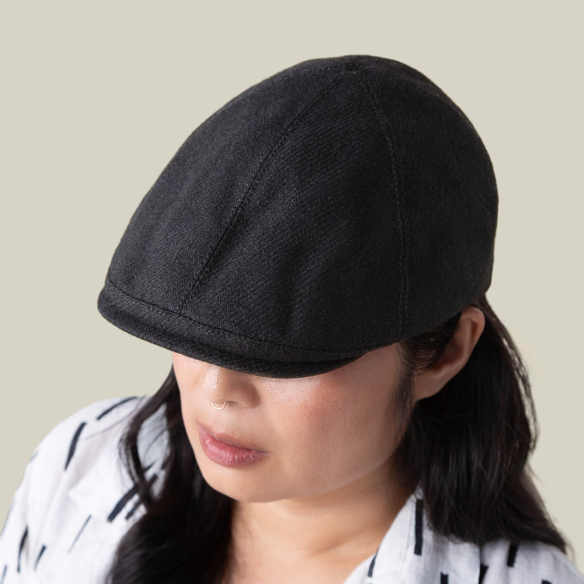 A person wearing the Goorin Bros. Ronald Amos black six-panel flat cap with a two-inch brim and a white patterned shirt, looking downward against a plain beige background.