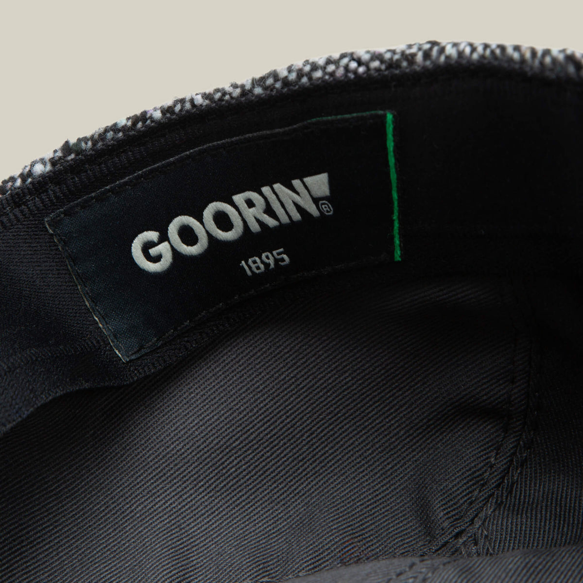 Close-up of the inside label on a grey tweed cap, showing the brand "Goorin Bros." and the product name "Gibbins" on a black fabric background—a classic piece for men's fashion.