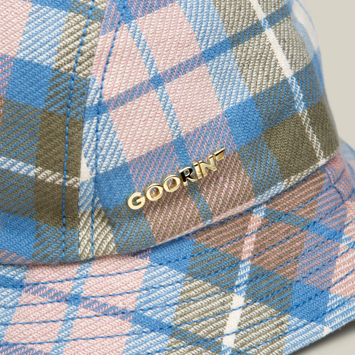 Close-up of the Bespoke Wordmark Bucket by Goorin Bros., a plaid bucket hat in blue, pink, brown, and olive green with a small gold GOORIN wordmark on the fabric.