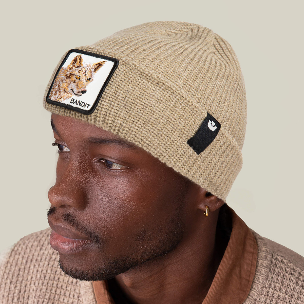 A man looks off to the side wearing The Bandit Beanie by Goorin Bros., an earth-tone knit winter hat featuring a coyote patch with the word "BANDIT" and a small black logo patch.