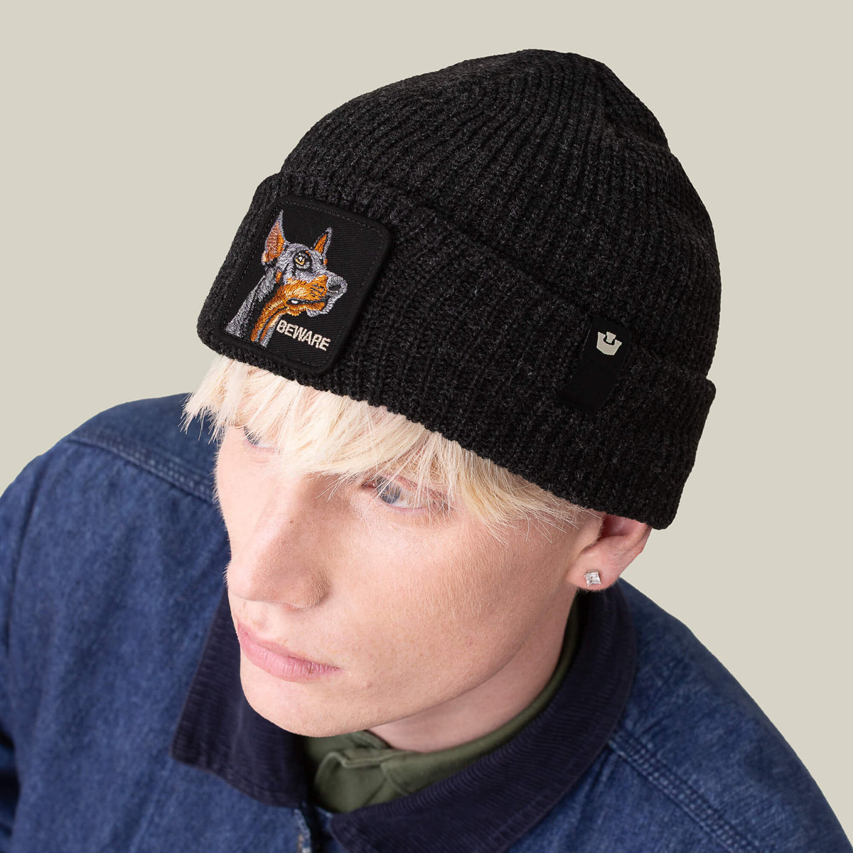 A person with blonde hair wears The Beware Beanie by Goorin Bros.—a heavyweight black knit beanie with a dog and "BEWARE" patch—styled with a denim jacket and stud earring.
