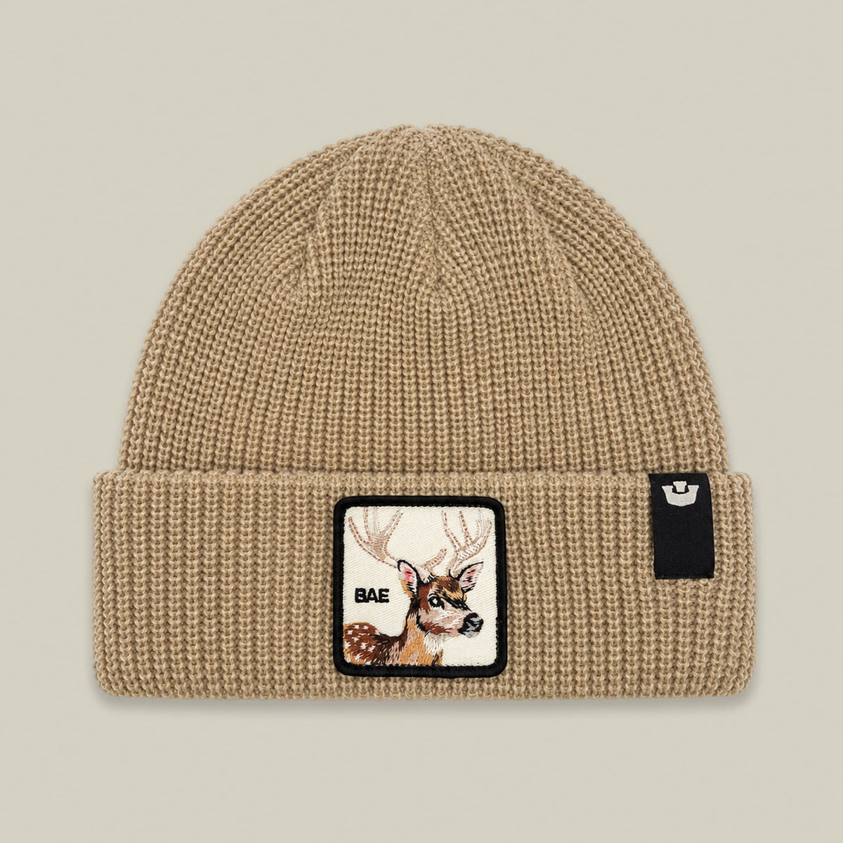 The Bae Beanie by Goorin Bros is a tan ribbed knit beanie featuring a woven patch with a deer graphic and "BAE" text on the front, displayed flat against a neutral background.
