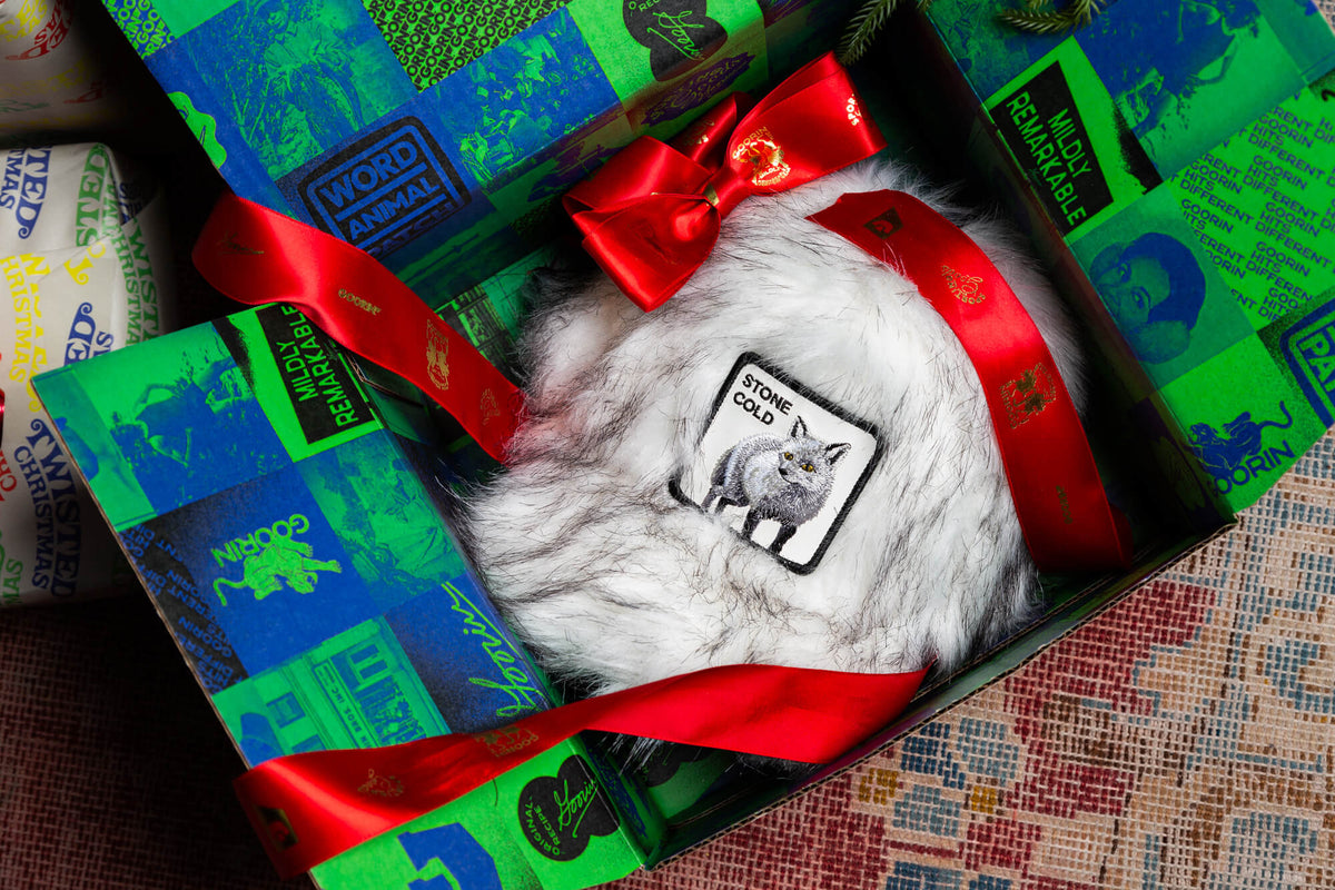 The Faux Fox by Goorin Bros. is a furry white faux fur hat featuring a Stone Cold wolf patch, elegantly presented in a gift box wrapped in green patterned paper and finished with a red ribbon bow—an ideal choice for those who love bold fashion accessories.