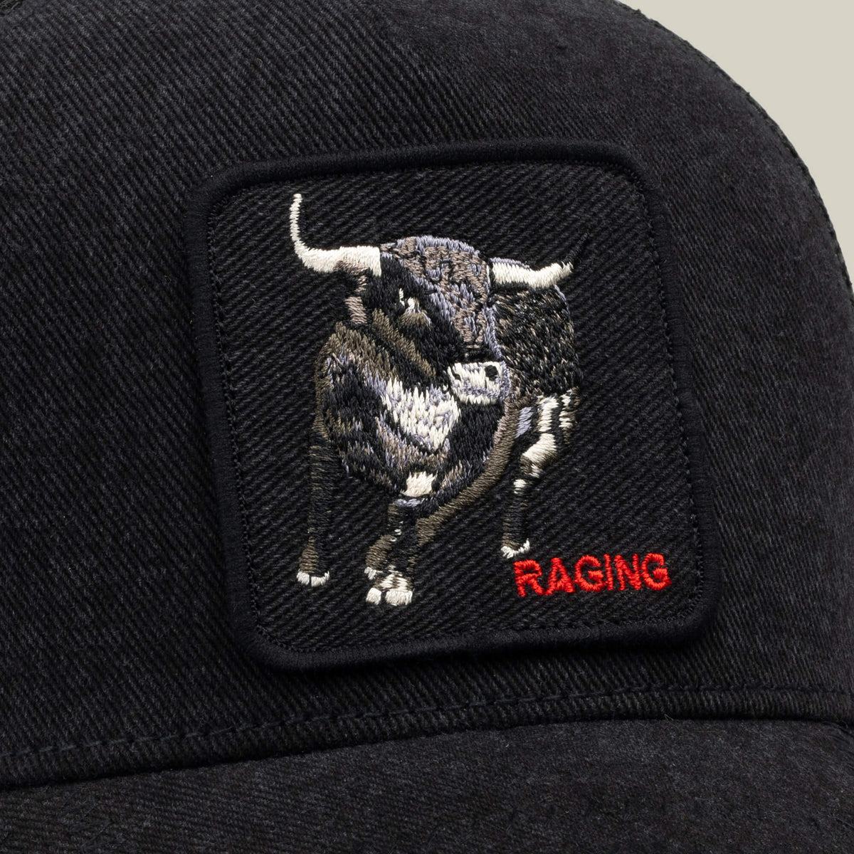 A close-up of the Goorin Bros. Denim Raging trucker hat, featuring black denim fabric, a stitched patch with a charging bull and “RAGING” embroidered in red, and breathable mesh panels for added comfort.