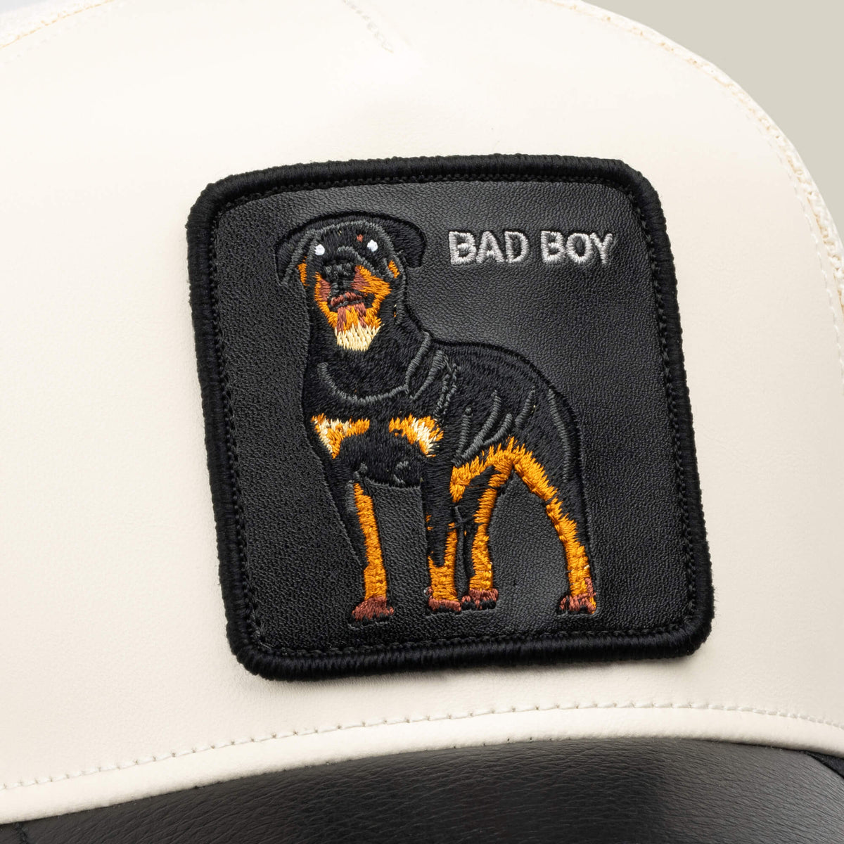 A white and black faux leather trucker hat from Goorin Bros., called the Faux Leather Rottweiler, features a stitched patch of a Rottweiler with the words BAD BOY on the front.